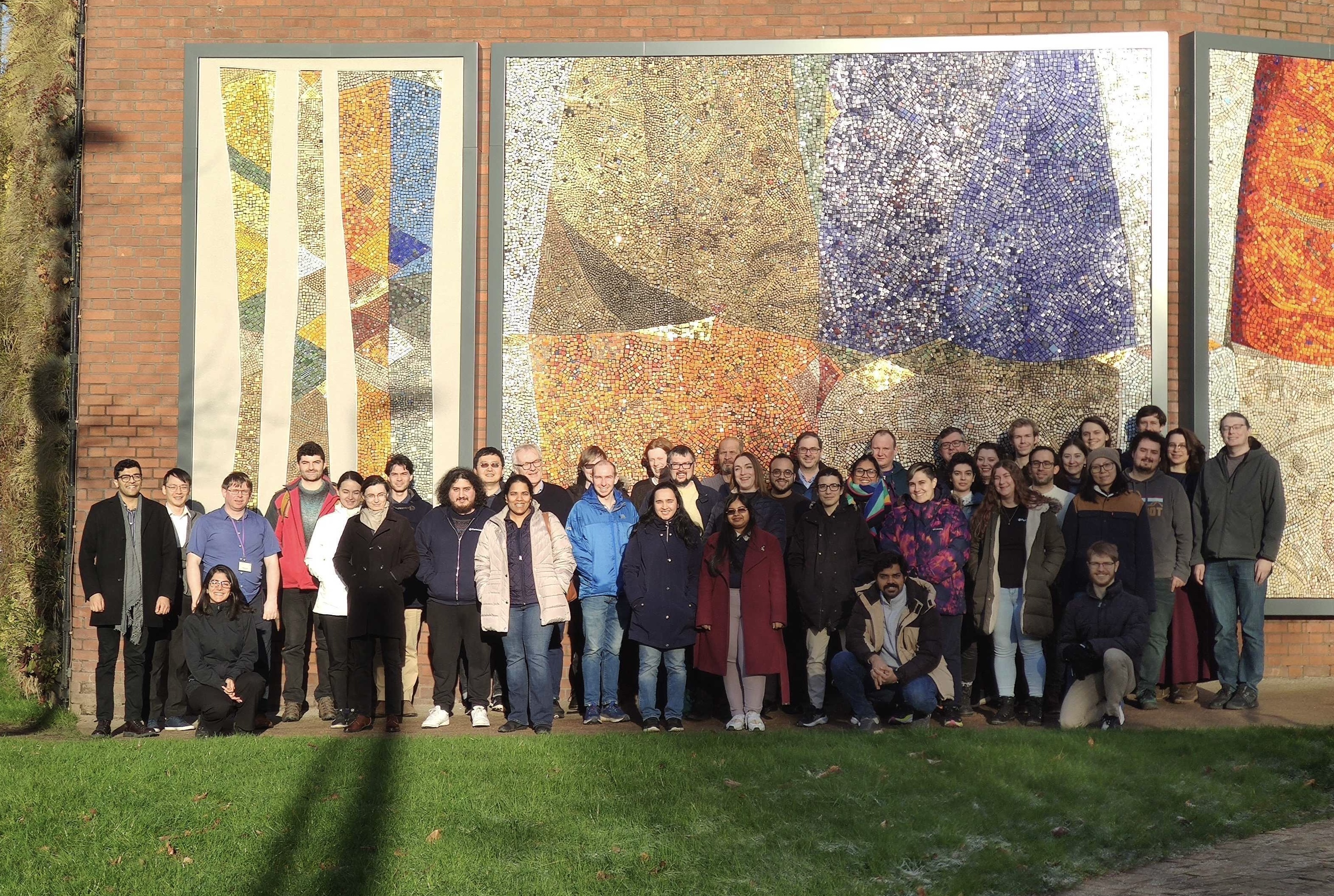 Members of the MicroBooNE Collaboration lined up in from of a decorative mosaic artwork on a red brick wall.