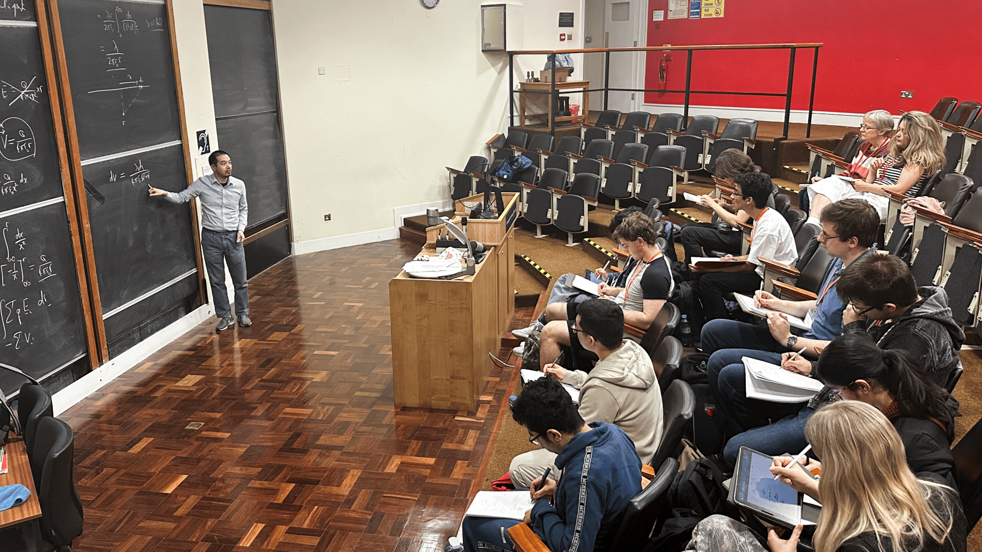 Lecture in classroom with students in seats and instructor at blackboard pointing at equations.