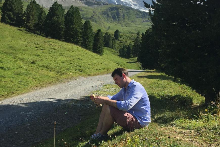 Daniel Hynds sitting down on the grass reading a book outside with mountains in the background