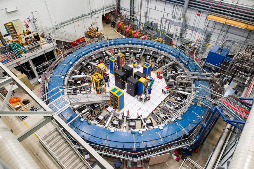 The Muon g-2 ring sits in its detector hall amidst electronics racks, the muon beamline, and other equipment.
