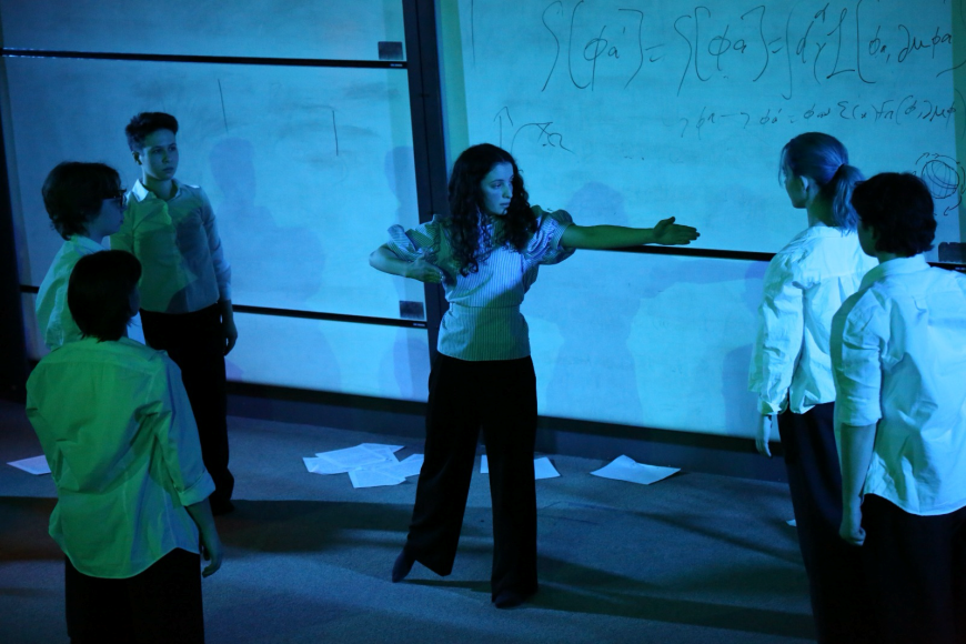 Image from play. Blue lighting with whiteboards in background. Actress in centre pointing to the right. 