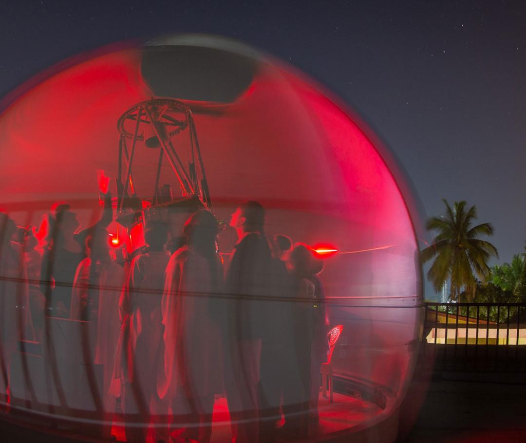 Female students in a red bubble with Professor Katherine Blundell looking up at the night sky through a telescope