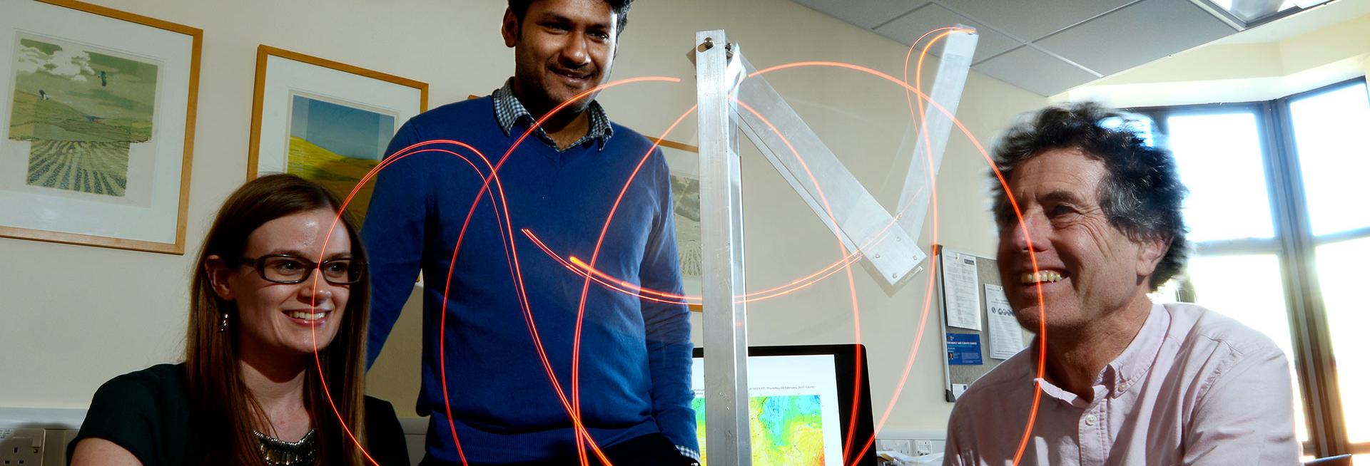 Image shows Prof Hannah Christensen, a male researcher and Prof Tim Palmer in an office, all smiling as they gaze at a device which is generating a pattern of curved orange lines of light. In the background is a computer screen with a heat map showing contours and coloured in the colours of the rainbow.