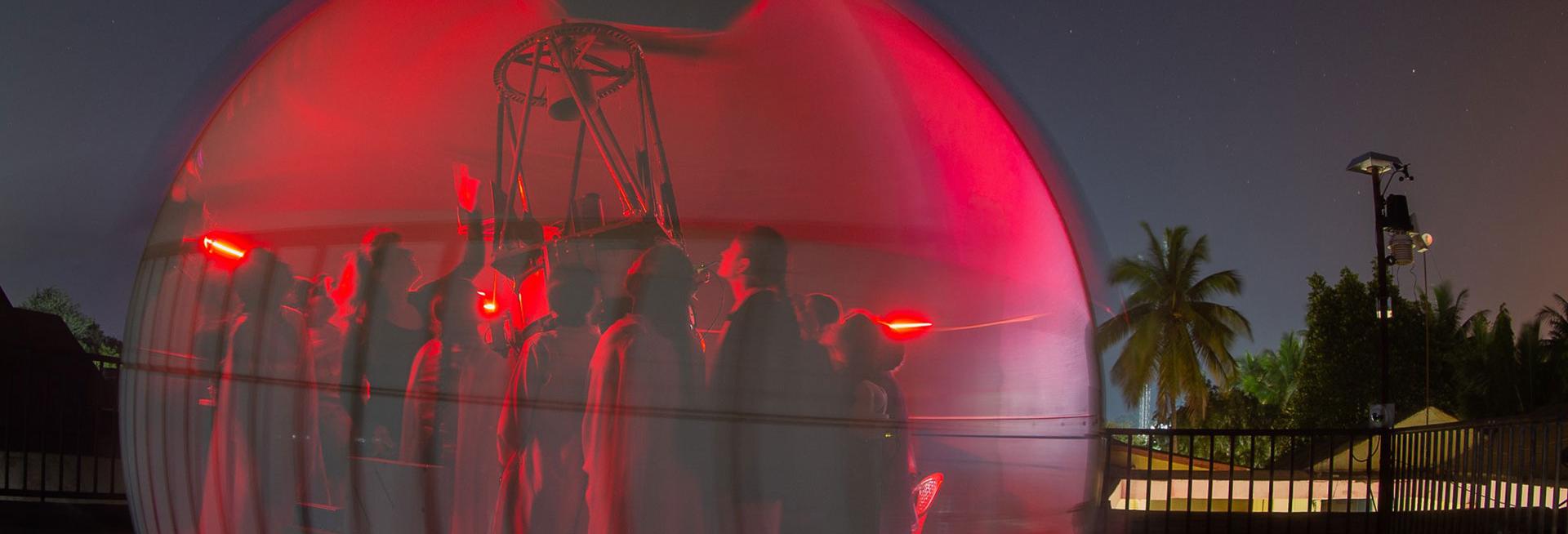 Female students in a red bubble with Professor Katherine Blundell looking up at the night sky through a telescope