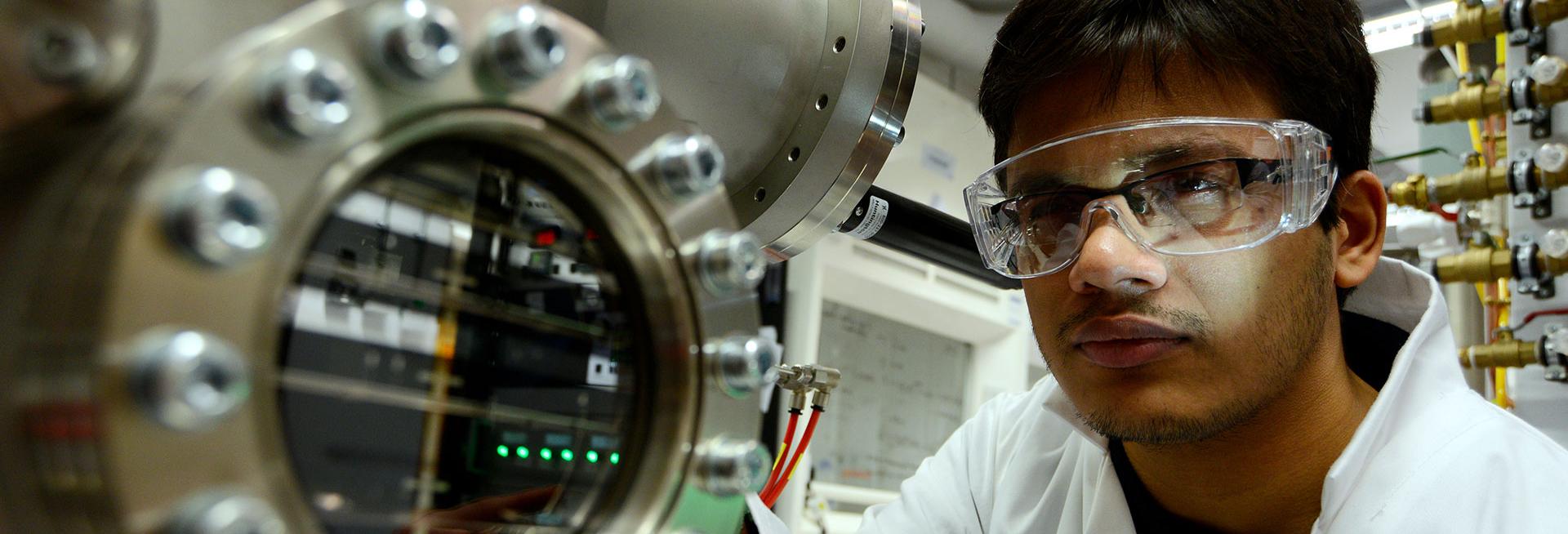 researcher wearing spectacles and safety glasses on top, and a white lab coat, looking intently at a large instrument made of silver metal. A row of bright green LEDs are visible through a clear window in the side of the instrument. There are fumehoods in the background.