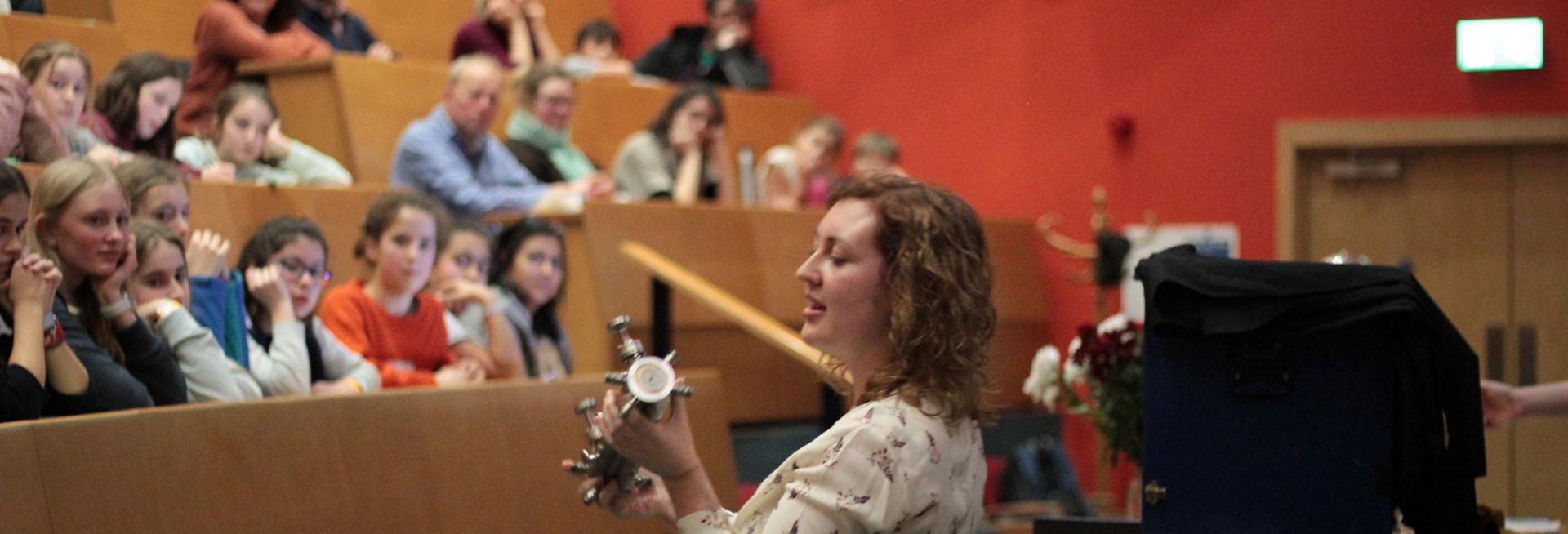 Female outreach officer holding an object talking to a lecture theatre of school students