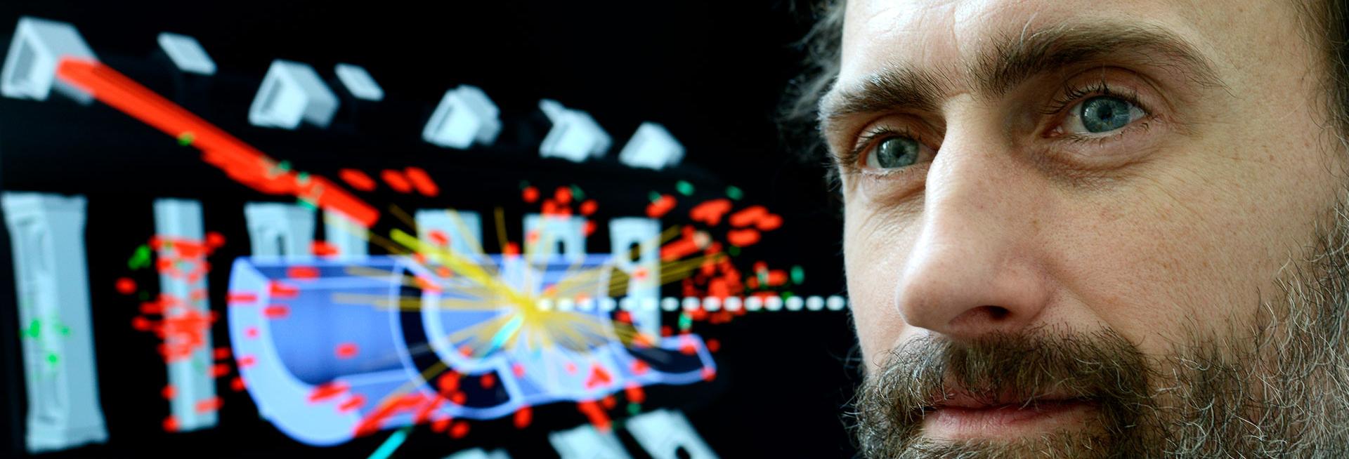 Profesor Chris Hays (in focus) and (out of focus) a diagram of the ATLAS detector which forms part of the LHC at CERN, with lines to show a particle collision and the resulting outputs of that collision.