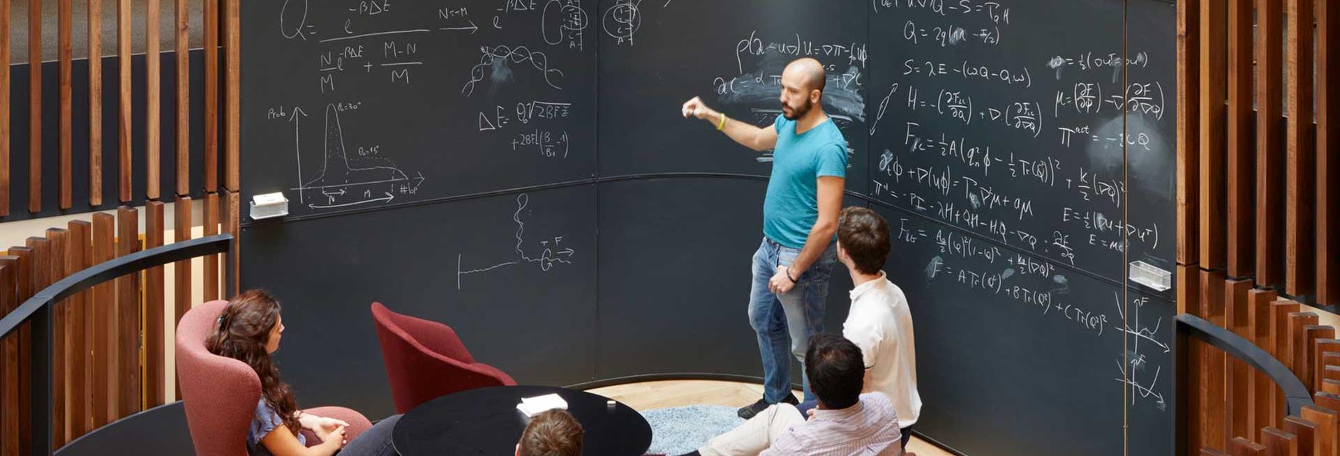 People sitting and standing around a blackboard discussing physics problems