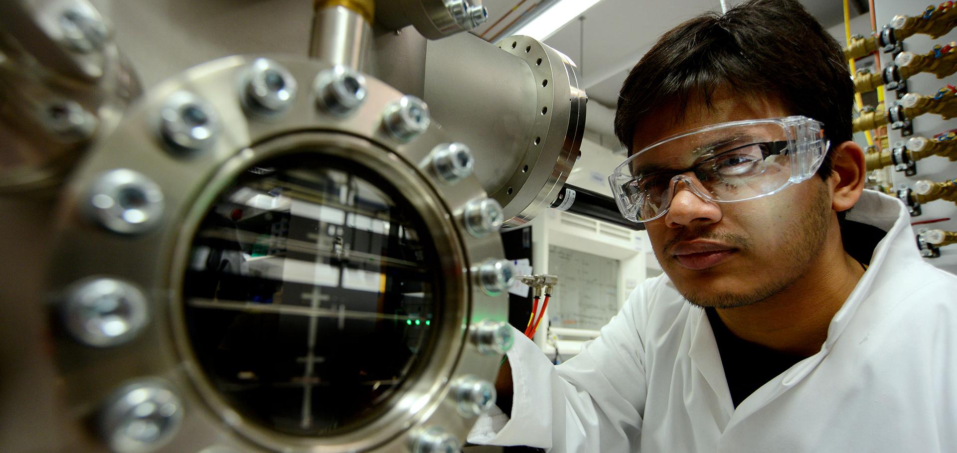 Man in white lab coat with protective eyewear working with scientific instrument.