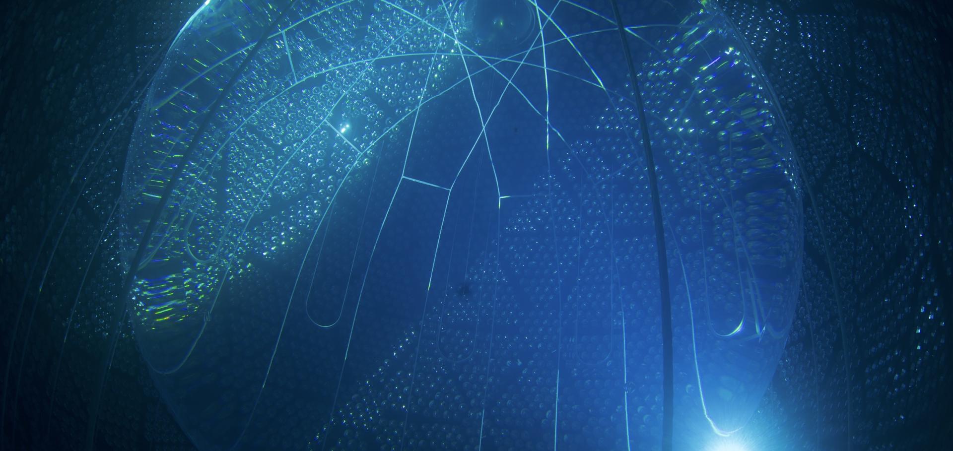 View from inside the filled SNO+ detector. A large transparent acrylic sphere filled with clear liquid, immersed in water. Surrounding the water, thousands of light-detecting photomultiplier tubes can be seen glinting with the reflective concentrators that surround them.